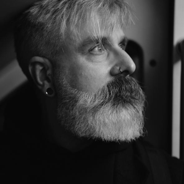 Black and white portrait of Adrian a bearded man with light hair, piercings, and a thoughtful expression, looking off to the side in soft lighting.